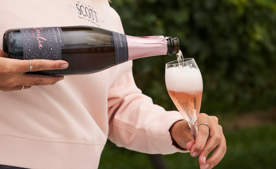 Woman pouring champagne into a glass outdoors with greenery in the background